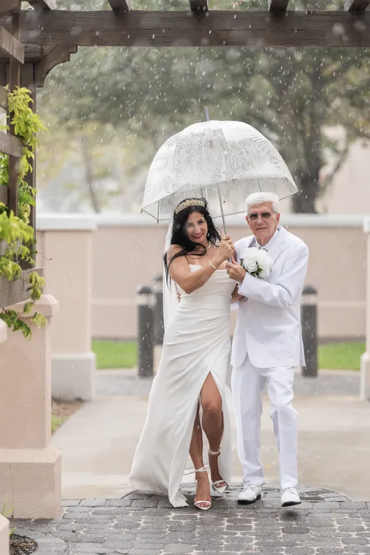 The couple walks beneath a wooden pergola with an umbrella during rain, their white outfits contrasting the grey weather.