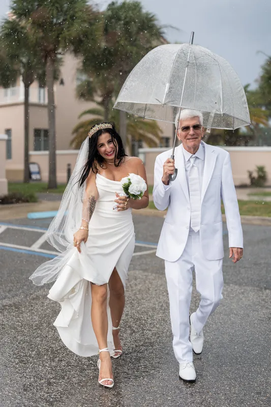 Mike and Isury walk together under a clear umbrella outside the courthouse, both in white attire with a white bouquet.