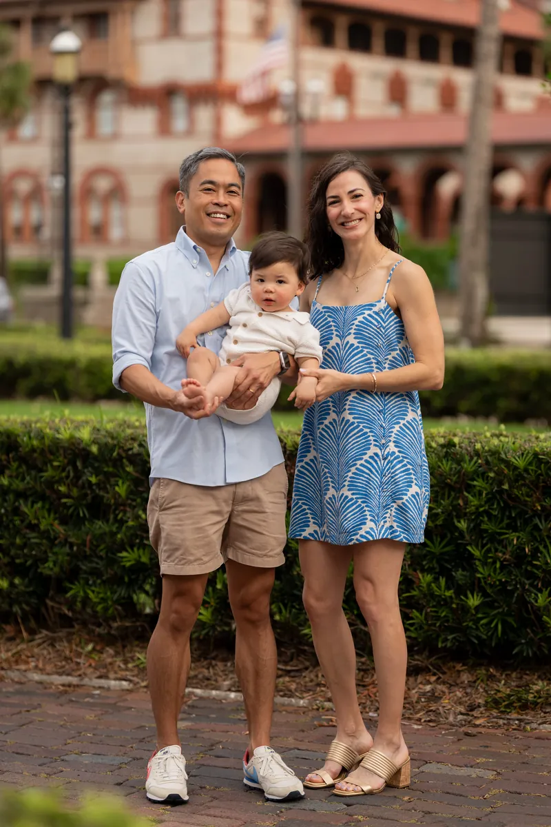 Joey and Monica hold their infant on a brick pathway with historic architecture framing the scene.
