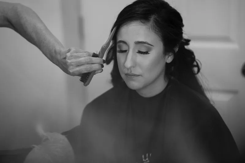 Monica sits in profile during hair preparation, eyes closed, while someone adjusts her dark hair at The Oldest House Museum.