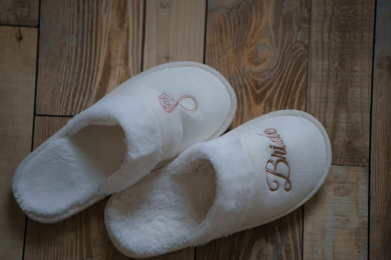 White embroidered slippers with "Mrs" monograms rest on rustic wooden flooring at the venue.