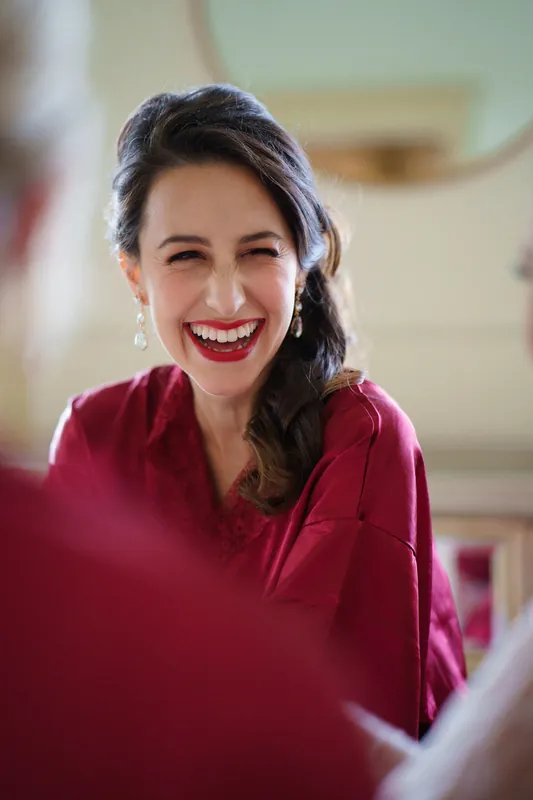 Monica laughs while wearing a burgundy shirt, her red lipstick and warm expression capturing a candid moment indoors.