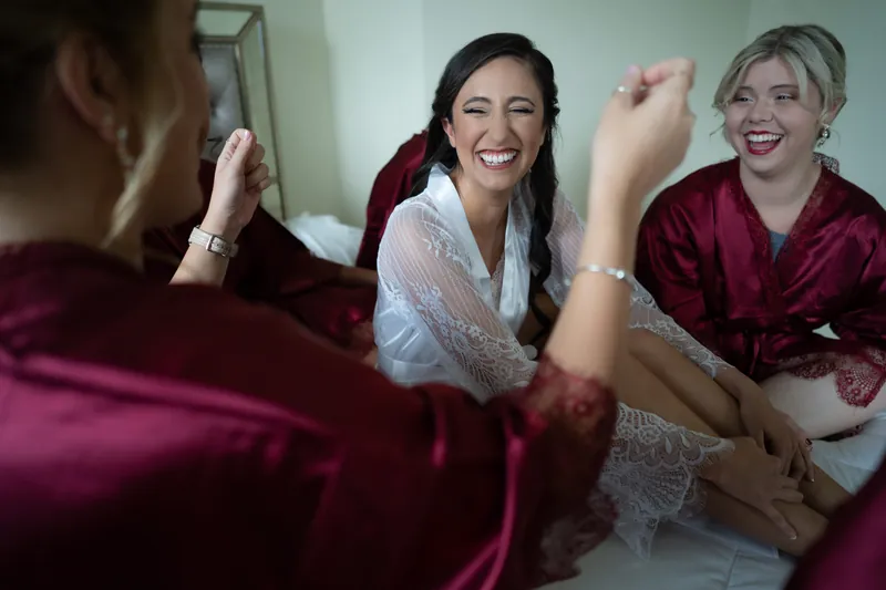 Monica laughs as she high-fives someone during getting-ready with her companions in burgundy robes.