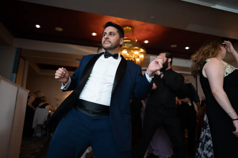 Michael dances energetically in a navy suit and bow tie, arms raised, surrounded by guests enjoying the celebration.