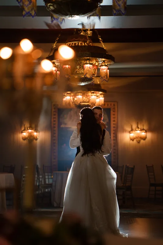 Monica stands alone in her white wedding dress, gazing at her reflection in a mirror framed by glowing sconces.