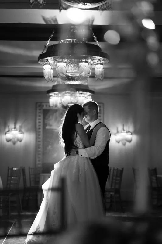 Monica and Michael embrace beneath an ornate chandelier in a black-and-white ballroom photograph.
