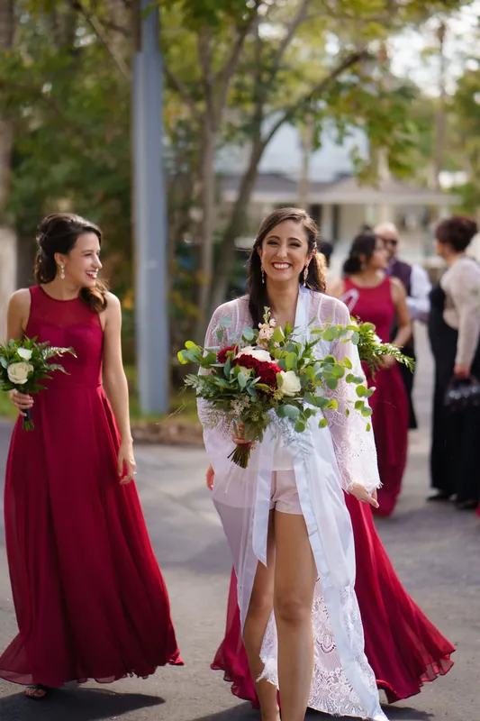 Monica walks with her bouquet toward the camera while bridesmaids in burgundy dresses follow behind on a tree-lined pathway.