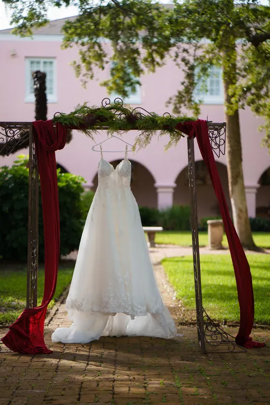 Monica's white lace wedding dress hangs from a wooden arbor draped in greenery and burgundy fabric in the courtyard.
