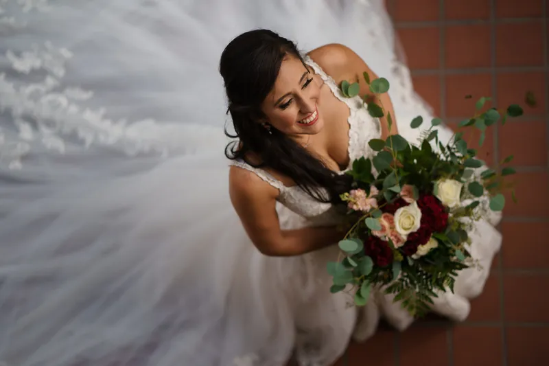 Monica holds a cascading bouquet of roses and greenery while smiling at the camera in a white lace gown.
