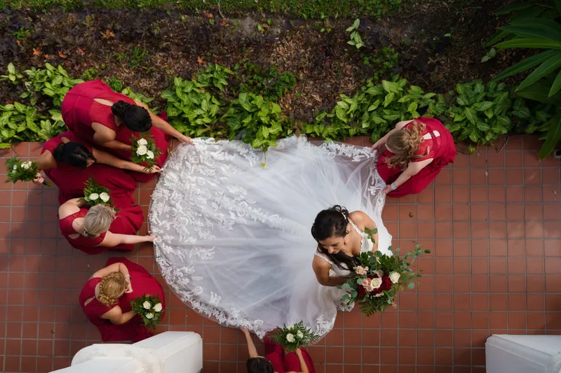 Monica in white lace stands on a brick courtyard surrounded by bridesmaids in burgundy holding white bouquets, shot from above.