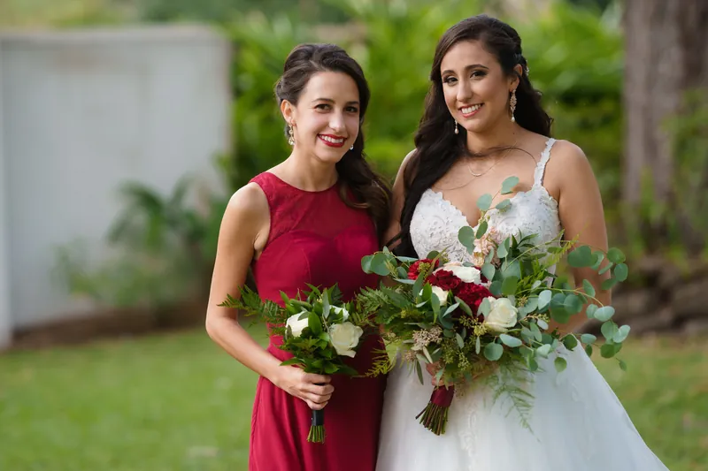 Monica and a bridesmaid in burgundy pose together holding mixed-flower arrangements with greenery in a garden setting.