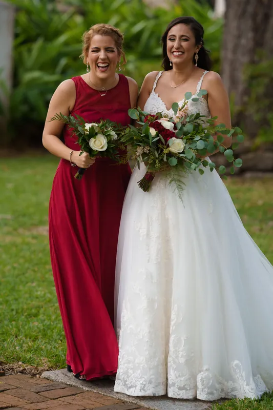 Monica and an attendant in matching burgundy and white gowns pose with their bouquets on the lawn, laughing together.