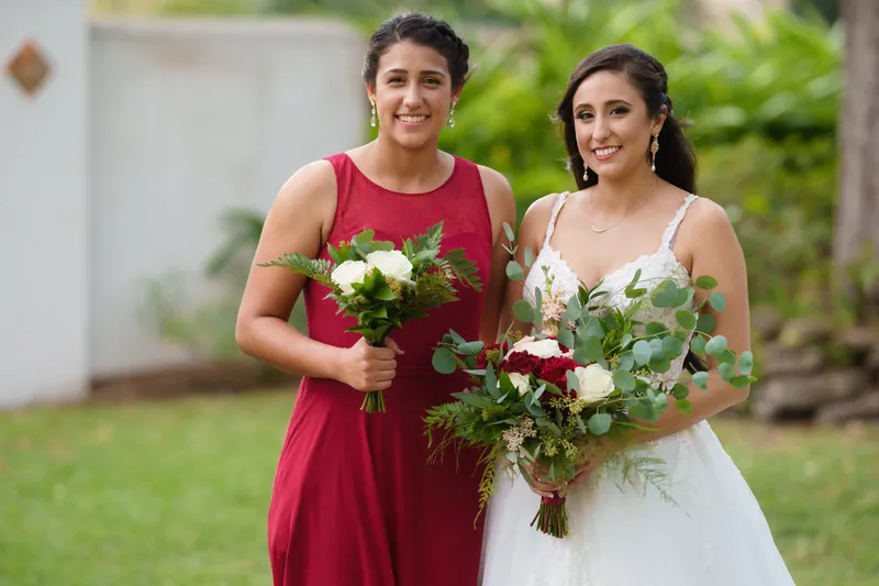 A bridesmaid in burgundy and Monica in white lace stand side by side holding white and greenery bouquets outdoors.