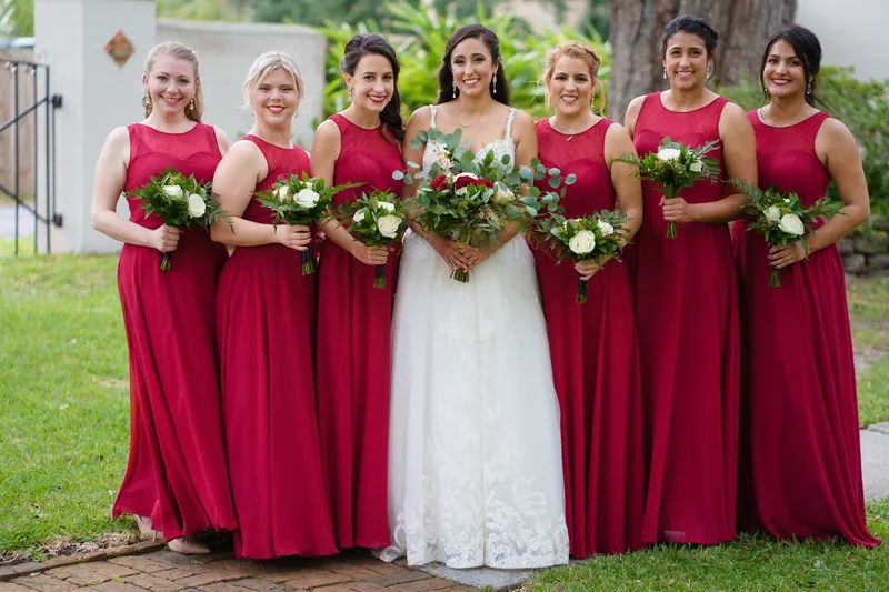 Monica in her white gown stands center with seven bridesmaids in deep burgundy dresses, all holding white flowers on the lawn.