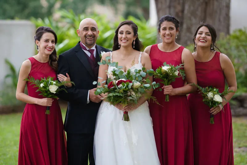 Monica stands with Michael and three bridesmaids in burgundy dresses, all holding white and greenery bouquets outdoors.