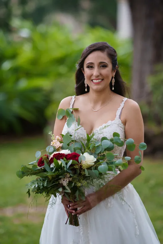 Monica holds a burgundy and white bouquet with greenery, smiling in her white sleeveless gown.