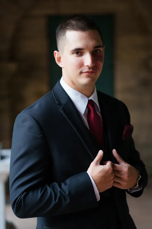Michael adjusts his navy jacket and burgundy tie in an indoor stone archway.