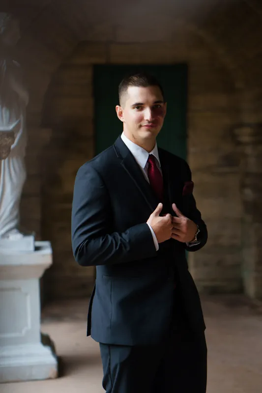 Michael stands in a navy suit with burgundy tie inside a historic stone corridor.