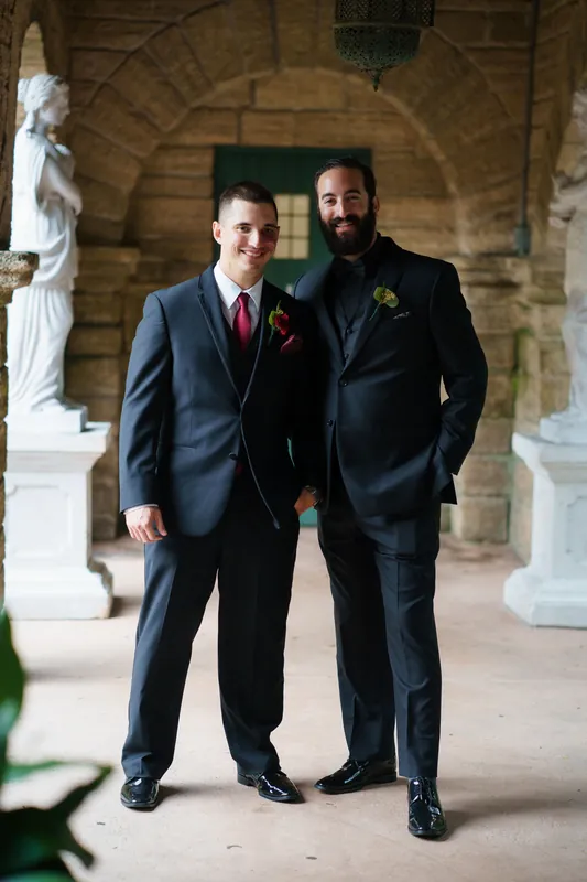 Michael and a groomsman stand together in navy suits with burgundy ties under a stone archway.