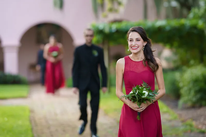 Monica holds her bouquet and beams in a burgundy dress as wedding guests gather behind her on the grounds.
