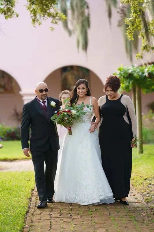 Monica stands between two family members on the brick pathway, holding her bouquet in her white embroidered wedding gown.