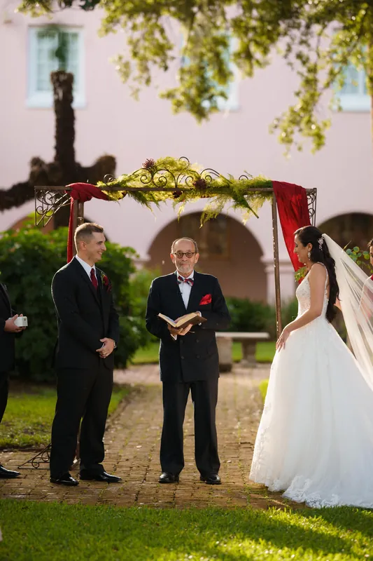 Monica and Michael exchange vows beneath a red arbor draped with greenery at The Oldest House Museum.