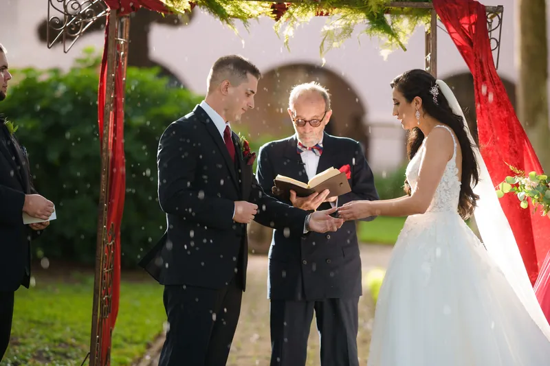 Confetti falls as the officiant reads vows while Monica and Michael hold hands under a red-draped arbor.