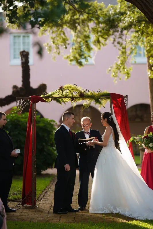 Monica and Michael clasp hands during their ceremony, the historic house framed behind the red-trimmed arch.