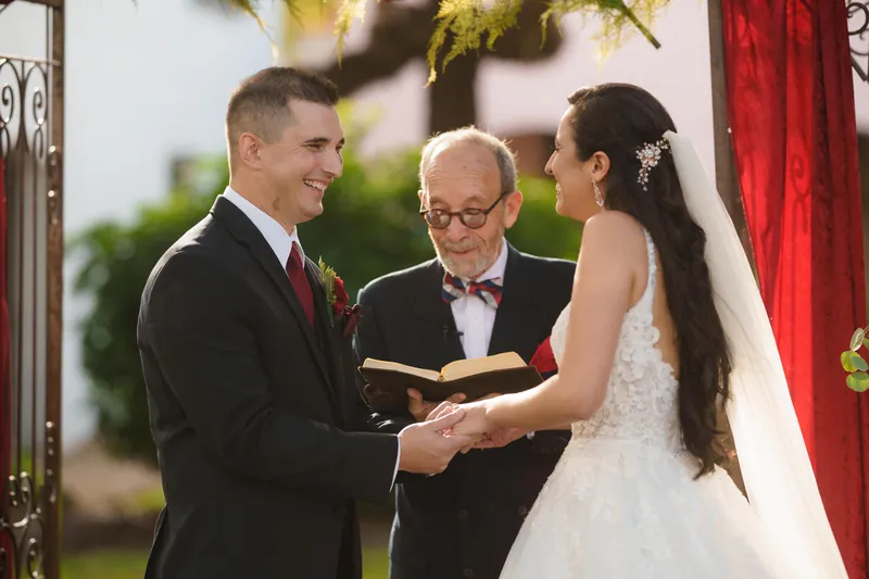 Michael smiles at Monica as their officiant leads them through vows, the couple framed by greenery and red ribbon.