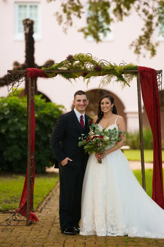 Monica and Michael stand under a red arbor with greenery, Monica holding a burgundy and white bouquet.