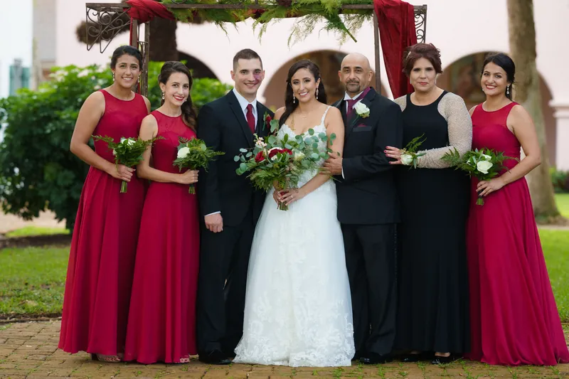 Monica stands center in white lace between bridesmaids in crimson and groomsmen in dark suits under the arbor.