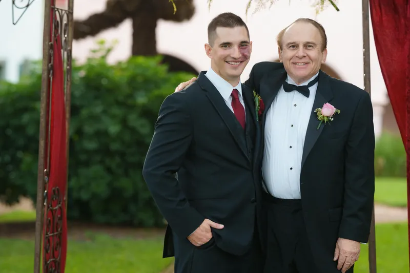 Michael smiles alongside a man in a tuxedo with a boutonniere, standing in front of the red arbor.