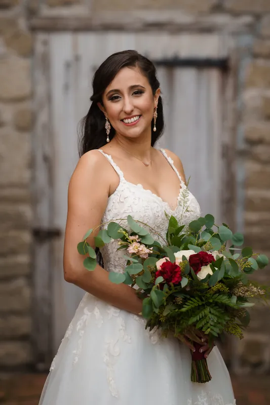 Monica in her sleeveless white gown holds a rich burgundy and green bouquet, smiling at the camera.
