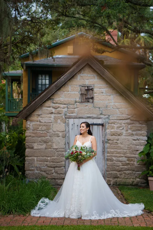 Monica stands in her flowing white wedding dress before the historic stone cottage, holding her bouquet.