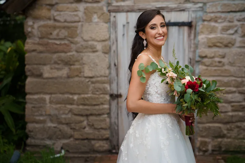 Monica holds a vibrant bouquet of roses and greenery, smiling over her shoulder against the historic stone backdrop.
