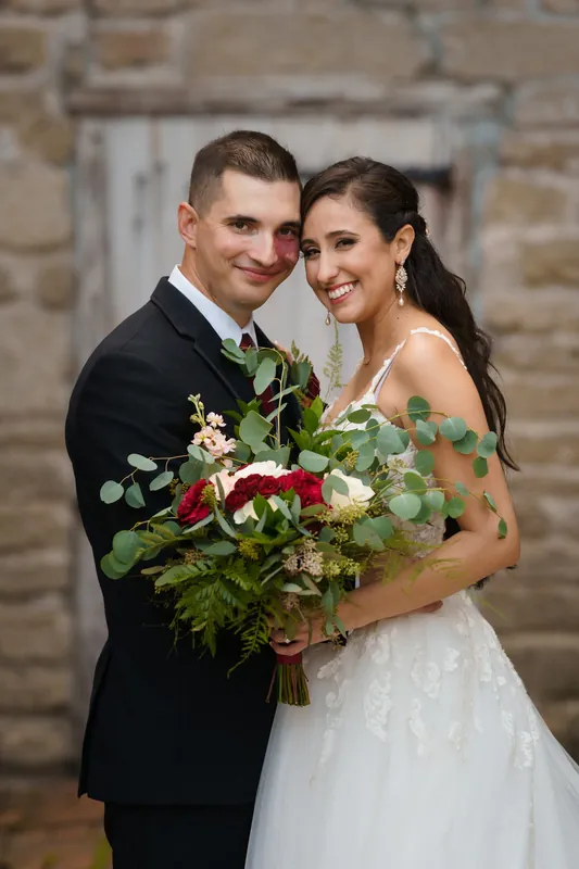 Monica and Michael pose together holding their lush bouquet, both beaming at the camera in front of the stone wall.