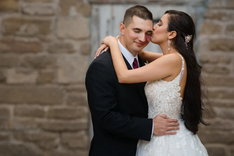 Monica kisses Michael's cheek as they embrace against the stone exterior at The Oldest House Museum.