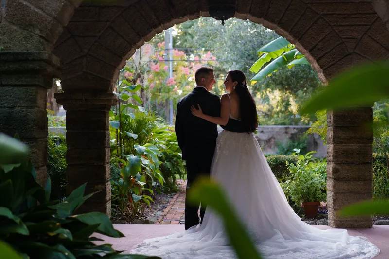 Monica and Michael kiss framed by a stone archway with lush greenery visible beyond at The Oldest House Museum.
