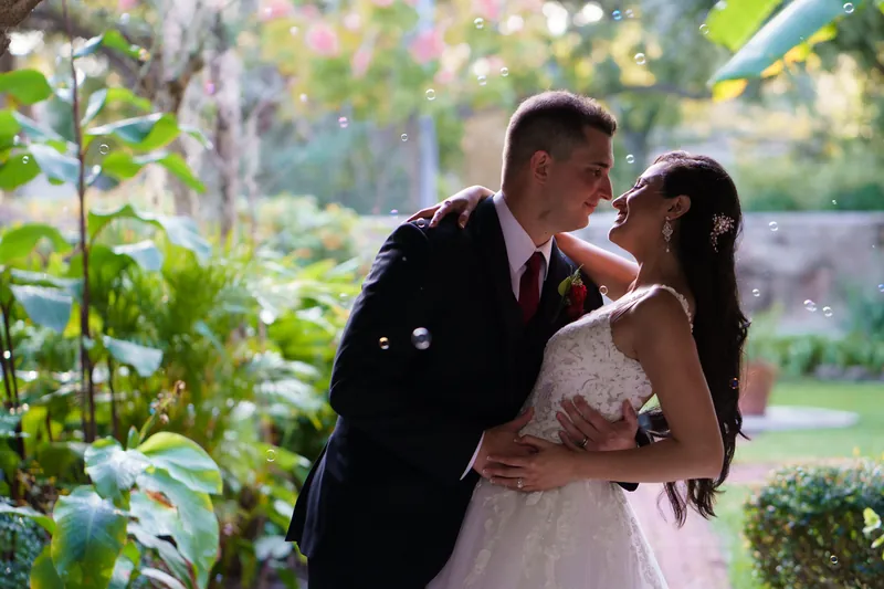 Michael leans in to kiss Monica's forehead as they stand surrounded by blooming flowers and greenery.