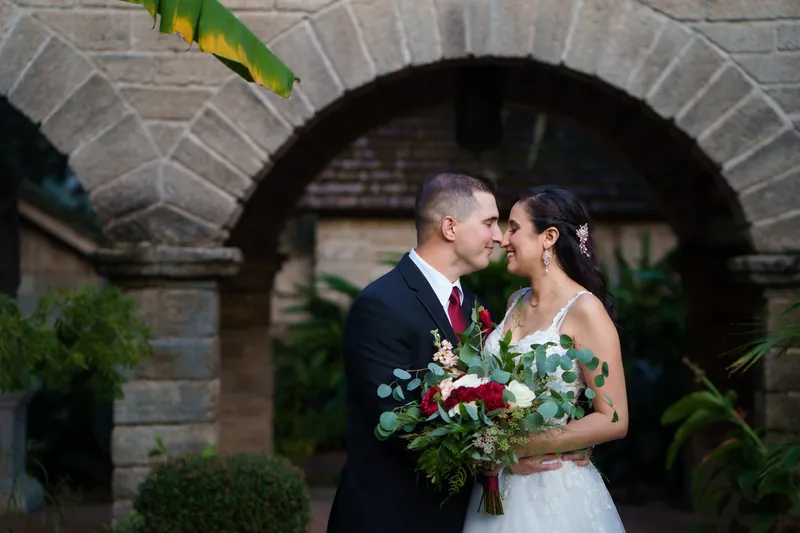 Monica and Michael kiss under a brick archway surrounded by tropical plants, holding a red and white bouquet.