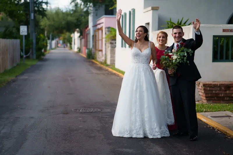 Monica and Michael wave from a brick street lined with historic buildings, with Michael holding the bouquet aloft.
