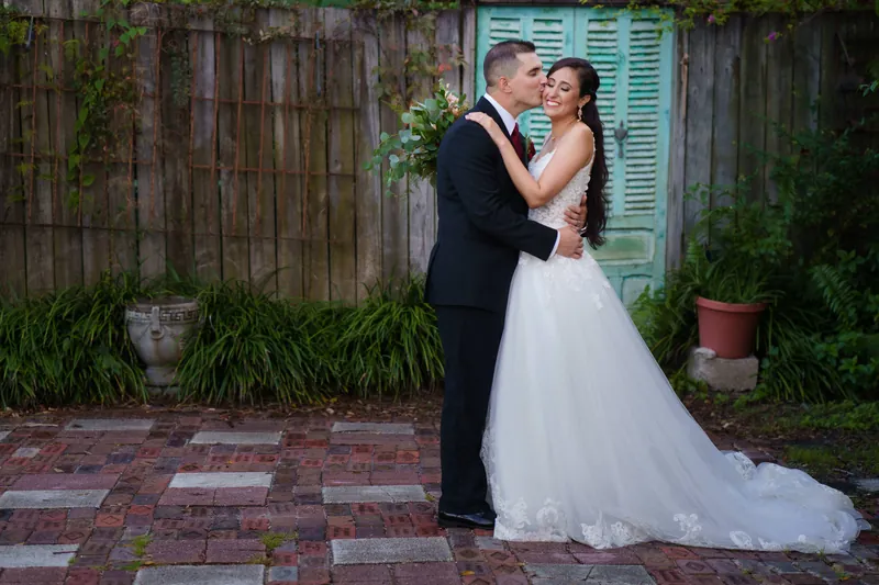 Michael and Monica embrace on a brick courtyard, with a weathered turquoise shutter and potted plants visible behind them.