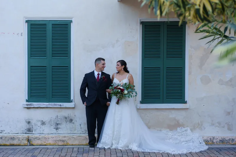 Monica and Michael walk past turquoise shutters of the whitewashed Oldest House Museum, Monica holding her bouquet.