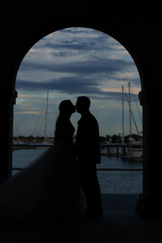 Monica and Michael kiss in silhouette beneath an arched passageway, with sailboats and water visible beyond.