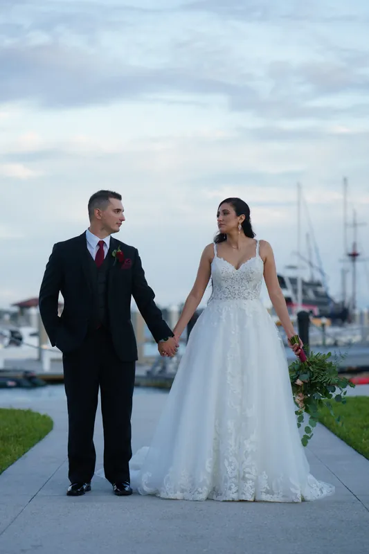 Monica and Michael hold hands while walking along a waterfront path with sailboats anchored in the harbor behind them.