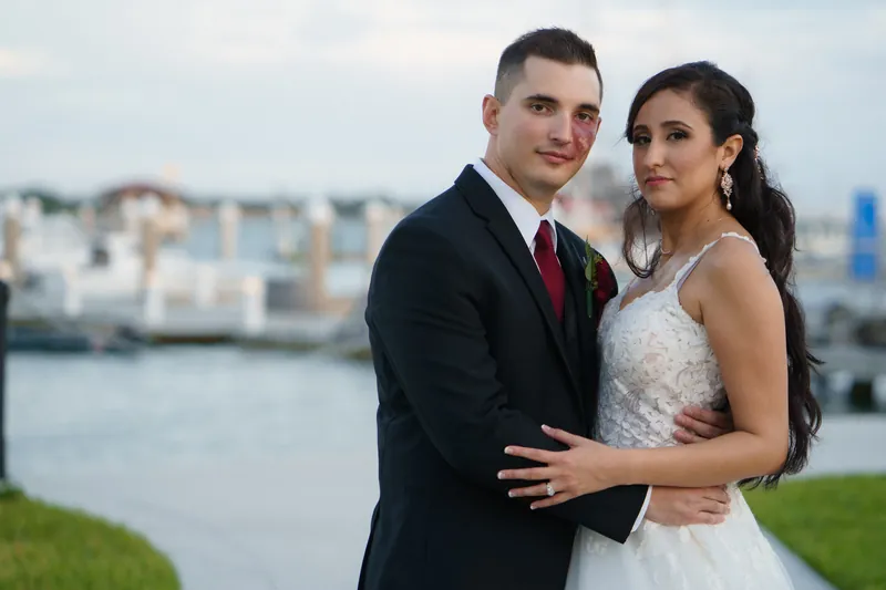 Michael embraces Monica from behind as they face the camera with the St. Augustine waterfront and boats in the distance.