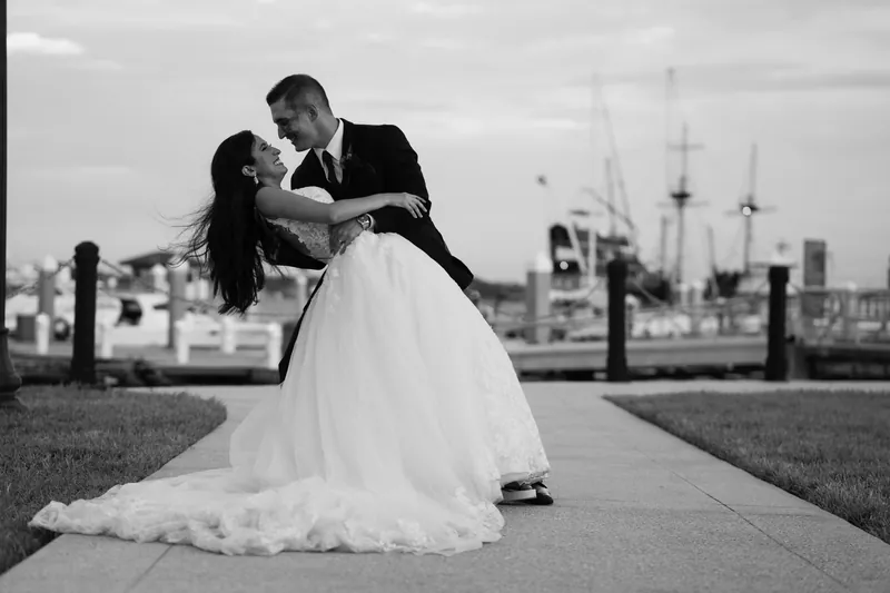 Michael dips Monica in a kiss on the waterfront promenade in black and white, with boats visible behind them.