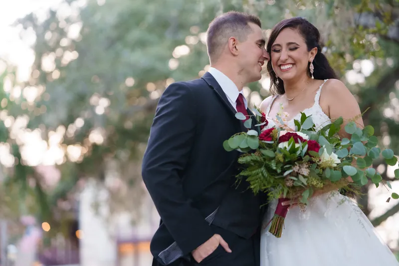 Michael kisses Monica's cheek as she smiles, both holding her rich burgundy and white bouquet beneath dappled sunlight.