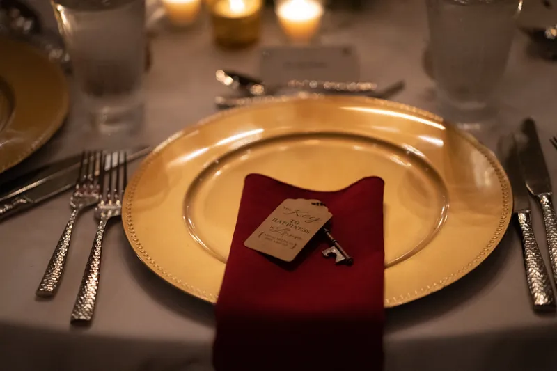 Gold charger plate topped with a burgundy napkin and place card at a formally set dinner table.