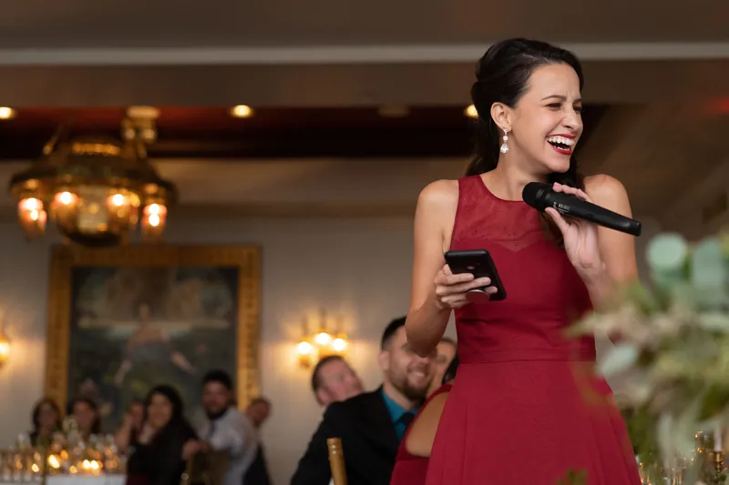 Woman in burgundy dress delivers a toast, smiling while holding a microphone in the elegant ballroom.